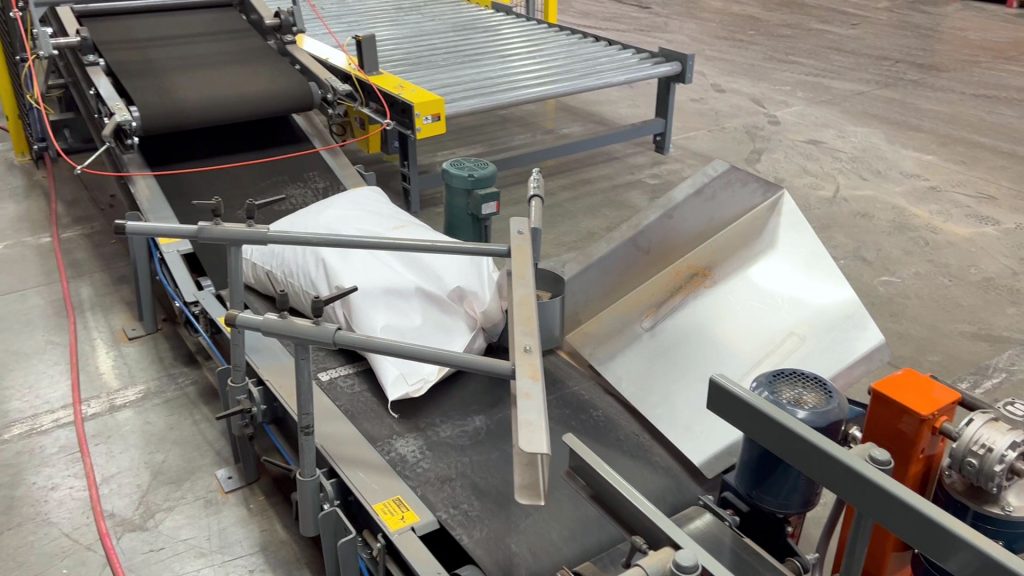 50# bag of nuts is knocked down before moving through a bag flattener conveyor